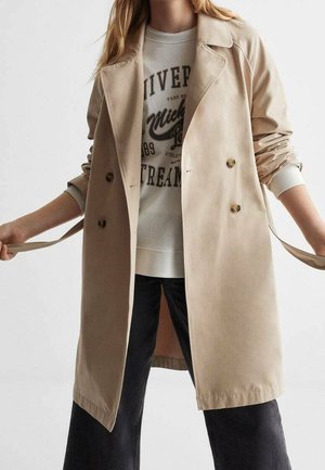 Femme portant un trench-coat beige croisé par-dessus un sweat-shirt blanc à motif graphique et un pantalon noir, tenant les passants de ceinture sur les côtés.