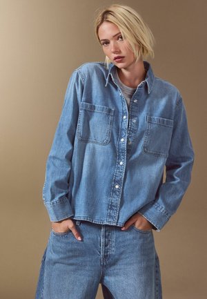 Młoda kobieta z krótkimi blond włosami, ubrana w za dużą niebieską kurtkę dżinsową i dopasowane jeansy, ręce w kieszeniach, na jednolitym beżowym tle.
