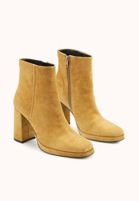 Bottes cheville en daim jaune moutarde avec un bout carré, un talon bloc et une fermeture éclair latérale. Texture lisse avec des détails de couture minimaux.
