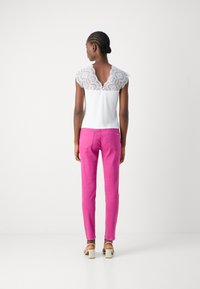 Haut blanc avec des manches capes en dentelle et un dos en forme de trou de serrure, associé à un pantalon ajusté rose vif et des sandales à talons carrés blanches.
