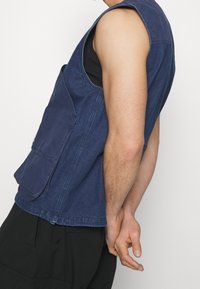Denimowa kamizelka bez rękawów w ciemnoniebieskim kolorze z kieszenią z przodu, o strukturalnym kształcie i kontrastowym ściegiem. Noszona na czarnej koszuli, widok z boku.