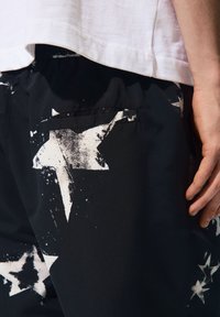 Čierne šortky s výraznou potlačou bielej hviezdy, elastickým pásom a bočnou vreckom. Materiál vyzerá hladko s jemne lesklou textúrou.