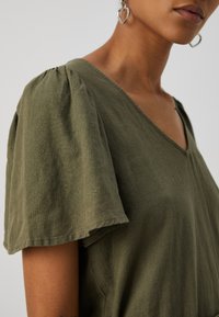 Top de algodão verde-oliva com decote em V, mangas curtas bufantes e um corte descontraído. Apresenta uma textura suave e detalhes de costura discretos.