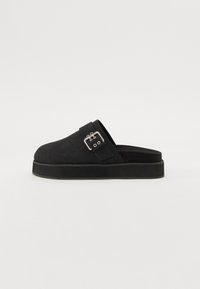 Crne klompe na slip-on od gamuza s debelim gumenim potplatom. Imaju jedan remen s srebrnom kopčom na gornjem dijelu za sigurno prianjanje.