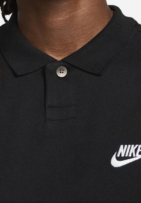 Zwarte poloshirt van textuurstof met een twee-knoopsluiting en een wit Nike-logo op de linkerkant van de borst.