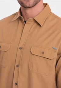Brązowa flanelowa koszula z kołnierzykiem na guziki, dwoma kieszeniami na piersi z patkami oraz kontrastującymi brązowymi guzikami. Miękka w dotyku o solidnej konstrukcji.
