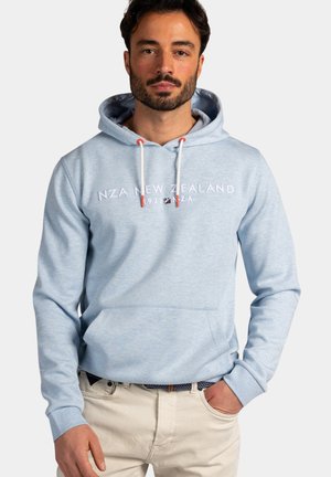 Man met baard die een lichtblauwe hoodie met de tekst "NZA New Zealand" en beige broek draagt, staand met één hand in de zak.