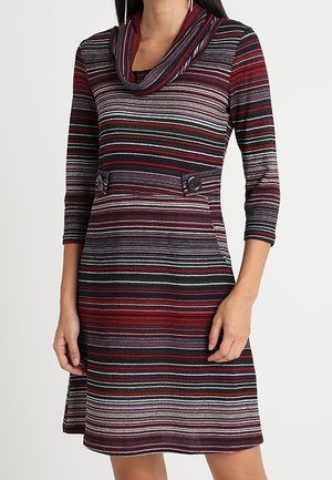 Kobieta ubrana w sukienkę sięgającą do kolan z poziomymi paskami w kolorach czerwonym, czarnym, szarym i białym, z rękawami 3/4, dekoltem typu łódkowego i ozdobnym zapięciem w talii na guziki.