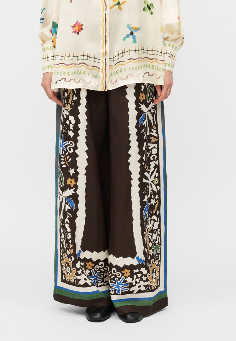 Καφέ παντελόνι με ευρύ τελείωμα, με λευκά ζιγκ-ζαγκ σύνορα και πολύχρωμα λουλουδάτα και αφηρημένα μοτίβα, συνδυασμένο με λευκό πουκάμισο με σχέδια.