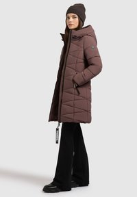 Pruun voodriga soe jakk kapuutsiga, küljetaskud, mustad tõmblukud ja logotägi, koos flöödidest mustade pükste ja jämedate mustade saapatega.