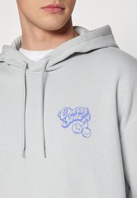 Felpa grigia chiara con logo frontale contenente il testo "Cherry Soda" in blu e ciliegie in stile cartone. Include un cappuccio con coulisse.
