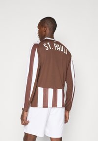 Dugmadžija s dugim rukavima smeđe boje sa bijelim prugama na rukavima, sa natpisom "ST. PAULI" u bijeloj boji na leđima, u kombinaciji s bijelim šortsem.