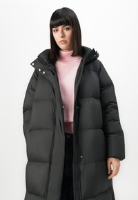 Donna con capelli neri lisci che indossa un cappotto imbottito nero sopra un maglione a collo alto rosa, in primo piano su uno sfondo bianco.