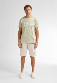 Homme se tenant face à l'objectif, portant un t-shirt vert clair de Petrol Industries, un short beige et des baskets blanches sur un fond blanc uni.