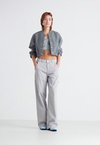 Chaqueta bomber gris corta con botones metálicos, combinada con pantalones anchos gris claro. El modelo lleva una camiseta gris sencilla y zapatos azules.