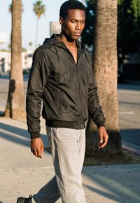 Jonge zwarte man in een zwart jack met ritssluiting en lichte broek lopend op een zonnig trottoir vlak bij palmbomen.