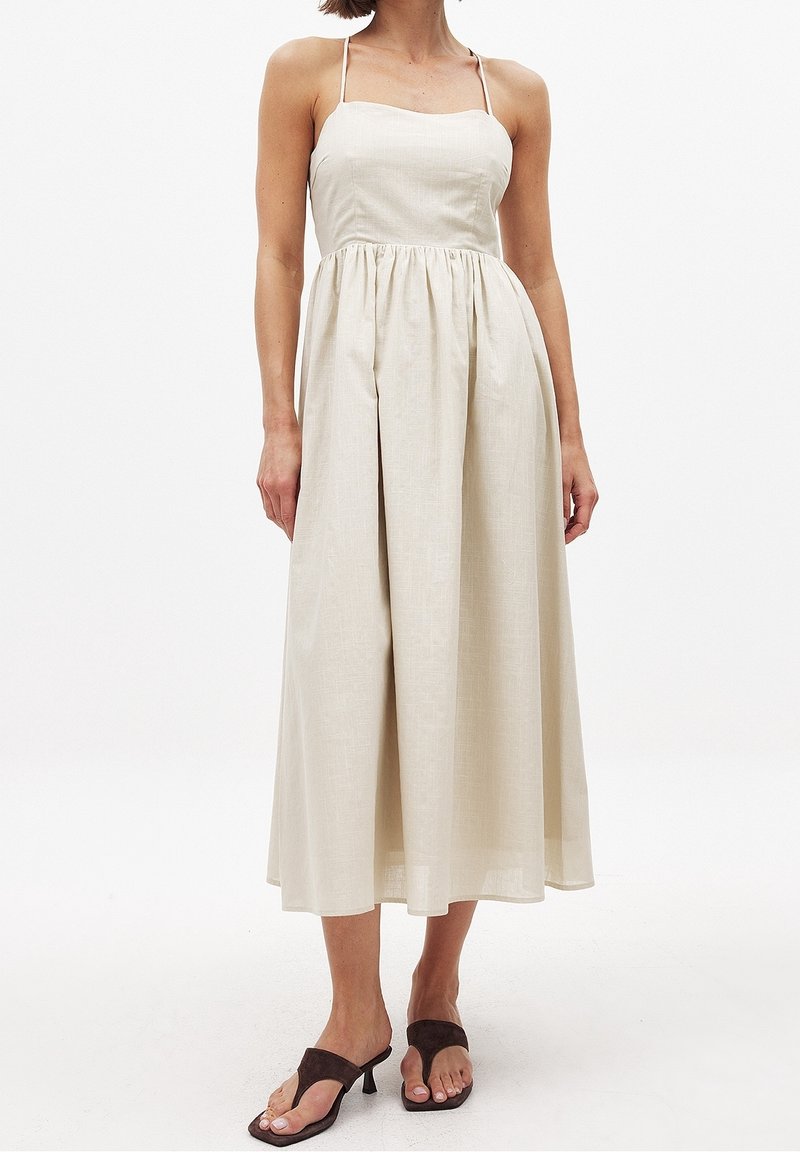 Robe en lin crème clair avec un corsage ajusté, une jupe plissée et des bretelles fines. Le tissu a une texture subtile et tombe en dessous du genou.