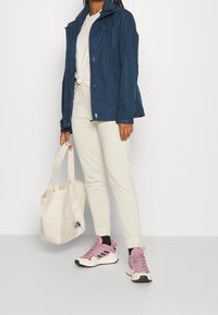 Personne portant une veste bleu marine, une chemise blanche nouée, un pantalon de jogging crème et des baskets roses, tenant un sac fourre-tout beige avec un logo.