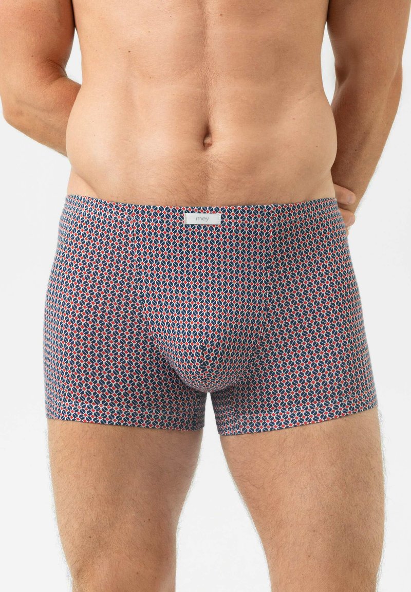Mann in gemusterten, eng anliegenden Boxershorts in Marineblau, Rot und Weiß, der mit den Händen hinter dem Rücken vor weißem Hintergrund steht.