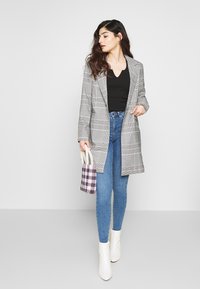 Blazer longo xadrez cinza, blusa preta, jeans skinny azuis, botas de tornozelo brancas, segurando uma pequena mala roxa e creme com padrão.