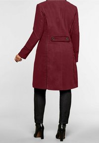 Abrigo burdeos con una silueta ajustada, detalles de doble botón en la cintura y costuras verticales, combinado con pantalones negros ajustados y tacones.