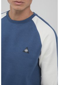 Blå sweatshirt med hvite raglan-ermer, rund hals og en liten svart trekantet logo-patch på brystet. Mykt bomullsmateriale.
