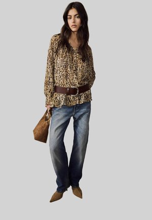 Junge Frau trägt eine Bluse mit Leopardenmuster, einen breiten braunen Gürtel, blaue Jeans, braune spitze Schuhe und hält eine braune Handtasche.