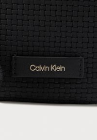Juodos tekstūruotos audinio medžiagos su juodu oda etikete, kuriame įspaustas sidabrinis "Calvin Klein" logotipas.