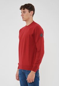 Maglione rosso a maglia con collo a giro e polsini a costine, con un patch logo sulla spalla sinistra. Indossato con jeans blu.