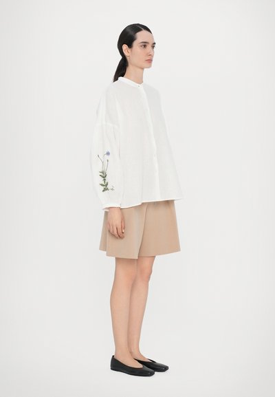Hvit linen skjorte med lange, puffete ermer og grønn og blå blomsterbroderi, kombinert med en beige skjørt og svarte ballerinasko.