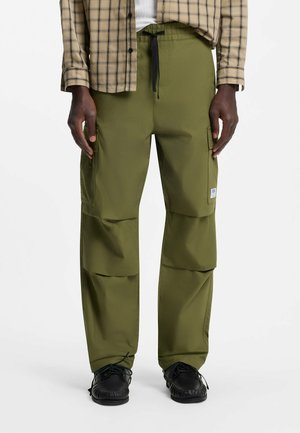 Uomo con pantaloni cargo verde oliva con tasche frontali e mocassini neri, in piedi davanti a uno sfondo semplice.