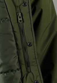 Grüne gepolsterte Jacke mit einem Reißverschluss, die einen strukturierten Stoff und schwarze Druckknöpfe aufweist, was ihr funktionales Design unterstreicht.