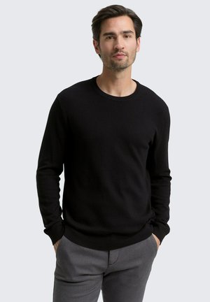 Homme aux cheveux foncés et barbate léger, portant un pull noir à manches longues et un pantalon gris, se tenant avec les mains dans les poches.