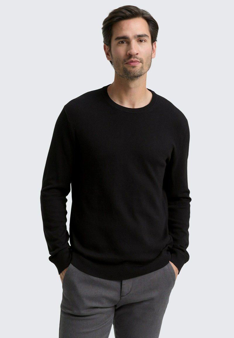 Mann mit dunklen Haaren und hellem Bart, der einen schwarzen Pullover mit langen Ärmeln und graue Hosen trägt und mit den Händen in den Taschen steht.