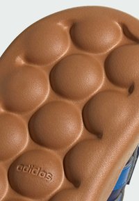 Semelle de chaussure en caoutchouc brun présentant une surface texturée avec des motifs circulaires en relief, un logo en relief, et une finition lisse sur les bords.