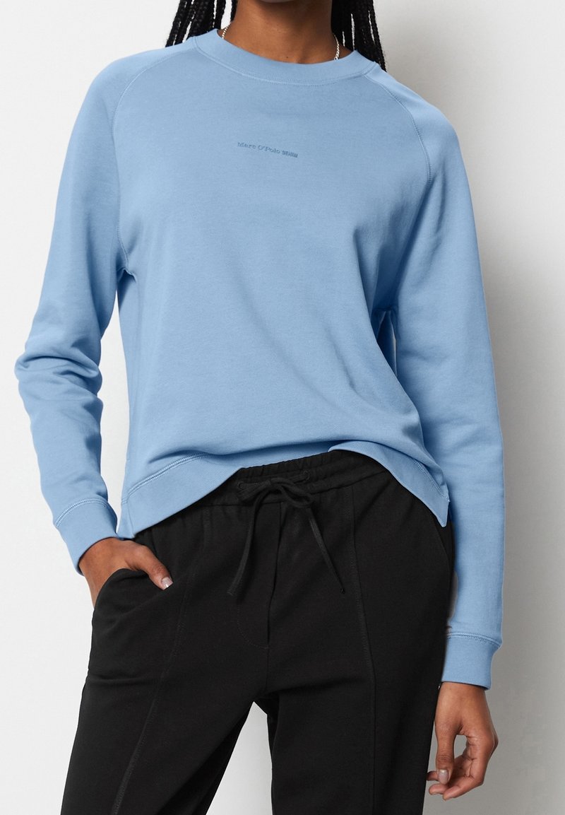 Sudadera azul claro con mangas largas, cuello redondo y un pequeño logo en el centro. Combinada con pantalones negros con cordón, que cuentan con bolsillos laterales.