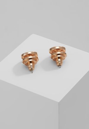 Par de pendientes de botón redondos en tono dorado con diseño circular en capas, descansando sobre un cubo blanco contra un fondo gris.