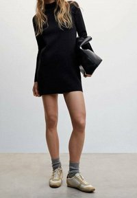 Robe noire à manches longues, silhouette ajustée, associée à des baskets blanches et beiges et à des chaussettes en tricot grises. Les accessoires comprennent un sac à main noir.