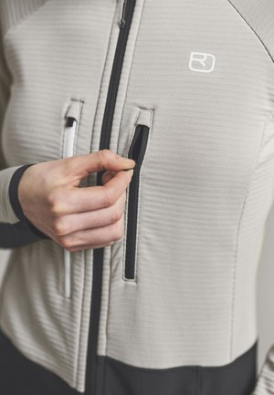 Hand hält Reißverschluss einer hellgrauen Jacke mit vertikaler schwarzer Reißverschlusstasche und weißem Logo auf der Brust.