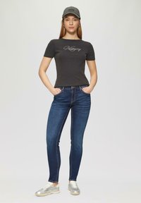 T-shirt noire à côtes avec logo en script, jeans slim-fit bleus et baskets argentées. Le modèle porte une casquette grise et pose les mains dans les poches.