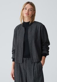 Grau strukturierte Bomberjacke mit Rundhalsausschnitt, gerippten Bündchen und Fronttaschen, kombiniert mit einem schwarzen Oberteil und passenden Hosen.