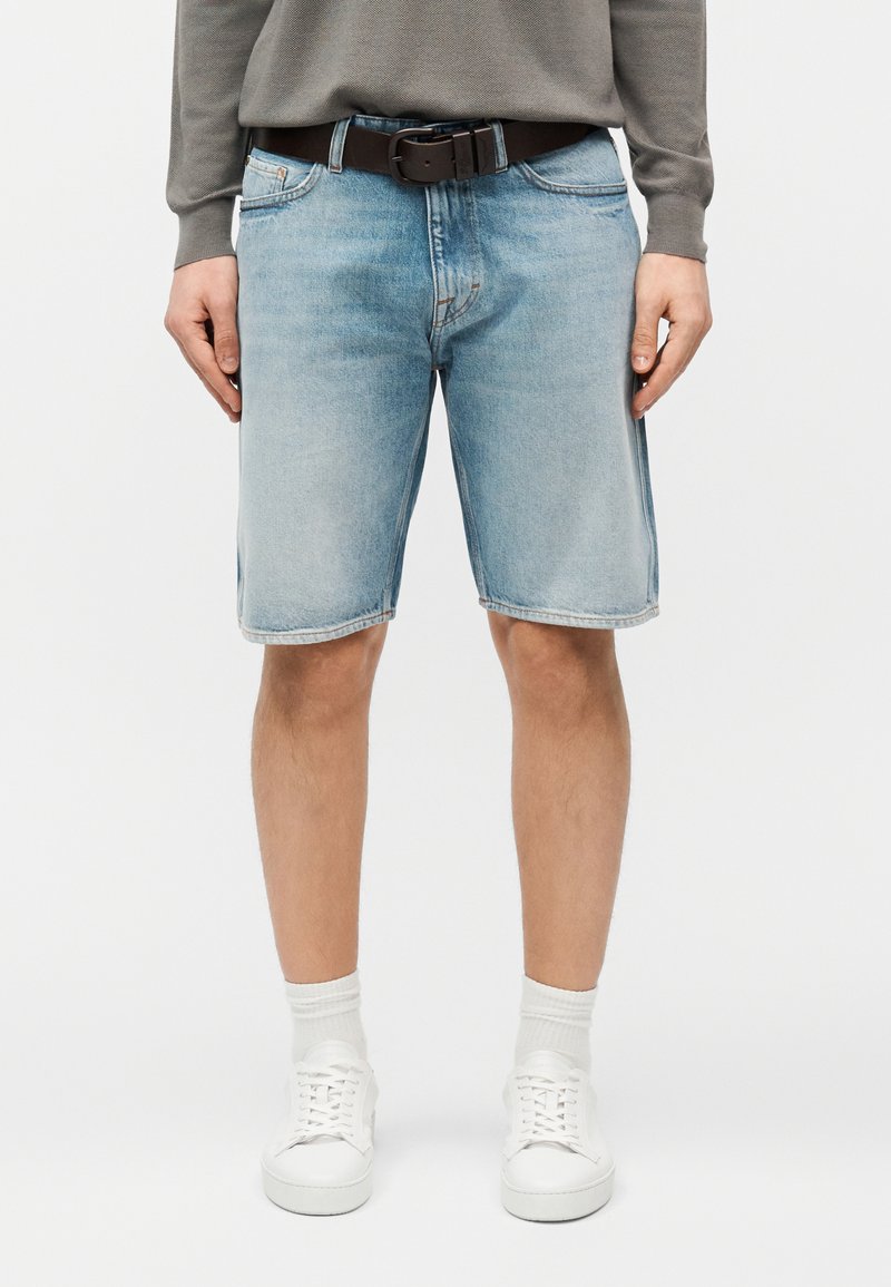 Άνδρας που φοράει ανοιχτό γαλάζιο τζιν σορτσάκι, λευκά αθλητικά παπούτσια, λευκές κάλτσες και γκρι μακρυμάνικο πουκάμισο με μαύρη ζώνη σε λευκό φόντο.