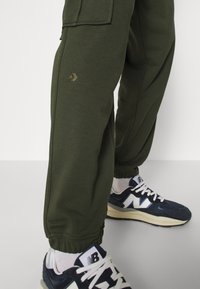 Olijfgroene cargojoggers met elastische boorden, voorzien van een zijzak en een klein gouden logo. Gecombineerd met marineblauwe en witte New Balance sneakers.