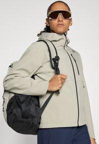 Giacca zip-up grigia chiara con superficie testurizzata, dotata di cappuccio e dettagli neri. Zaino in rete nera con spallacci regolabili. Occhiali da sole grandi.