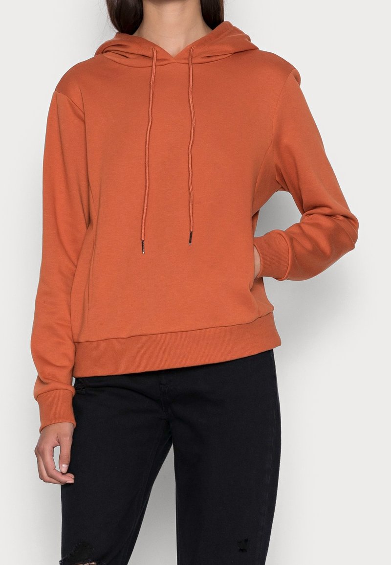 Kvinna som bär en rostfärgad huva med snörning och svarta slitna jeans, stående mot en enfärgad ljus bakgrund.