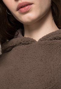 Brauner Fleece-Hoodie mit weicher Textur, ausgestattet mit einer abgerundeten Kapuze und einem dezenten Nahtdetail entlang des Halsausschnitts.