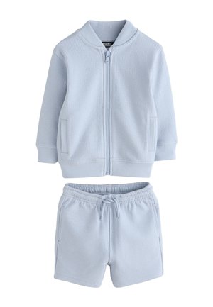 Kind trägt hellblaue, strukturierte Shorts und Jacke mit weißen Turnschuhen, steht auf einem beigen Boden vor neutralem Hintergrund.