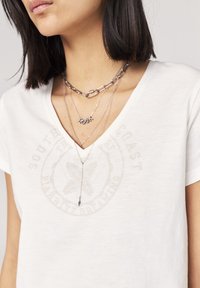 Camiseta blanca con cuello en V y un logo circular desvanecido. Collares de plata en capas; uno presenta letras "ROCK" y los otros tienen diferentes estilos de cadena.