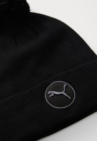 Berretto di maglia nero con superficie testurizzata, presenta un logo circolare grigio con un design di un gatto saltante sul risvolto. La parte superiore è completata da un pompon.