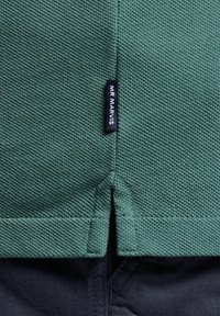 Tissu texturé vert avec un tissage fin, présentant une fente sur le côté à l'ourlet et une petite étiquette noire indiquant "MR MARVIS."